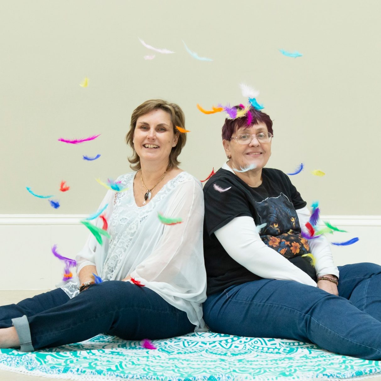 Two women sitting back to back on a colourful rug, smiling with confetti around them.